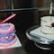 Two cakes on a bench, left cake is a heart shape iced with purple and pink colours, right cake is circular half iced with white